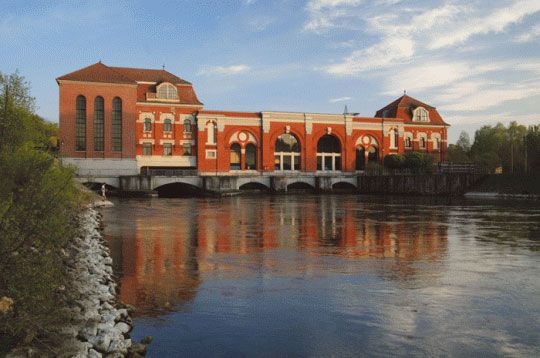 Rotes Backstein-Wasserkraftwerk an einem ruhigen Fluss unter blauem Himmel.