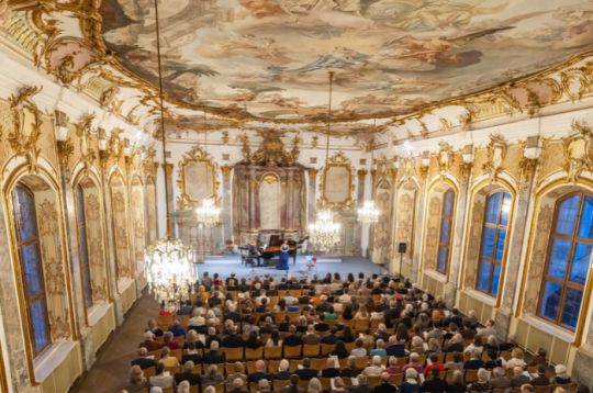 Eleganter Konzertsaal mit Publikum, verzierten Kronleuchtern und bemalter Decke.