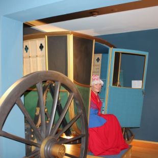 Eine Person in historischer Kleidung sitzt in einer verzierten, vintage Kutsche, die in einem Museum ausgestellt ist.