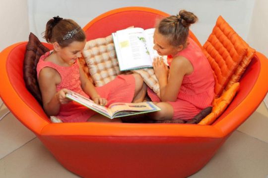 Zwei Mädchen in rosa Kleidern lesen Bücher auf einem orangefarbenen Sitz mit Kissen.