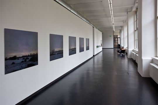 Un lungo corridoio con opere incorniciate su una parete bianca e grandi finestre sulla destra, con un pavimento scuro.