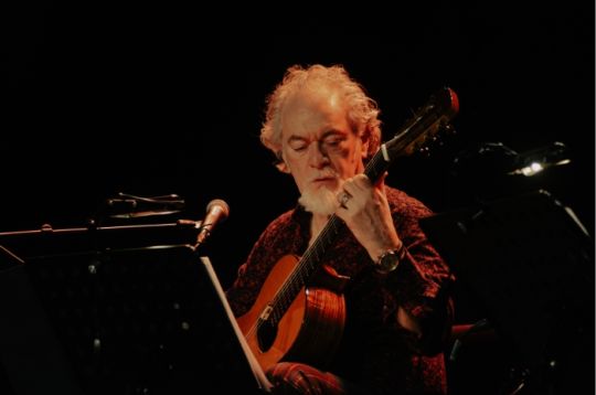 Ein Musiker mit weißen Haaren spielt auf der Bühne Gitarre, von einem Scheinwerfer beleuchtet.