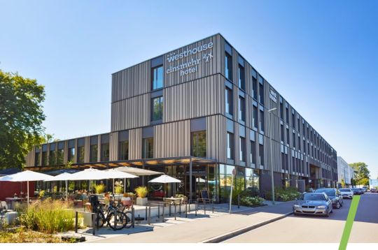 Modernes Hotelgebäude mit Fahrrädern und Außensitzplätzen unter Sonnenschirmen, vor klarem blauen Himmel.