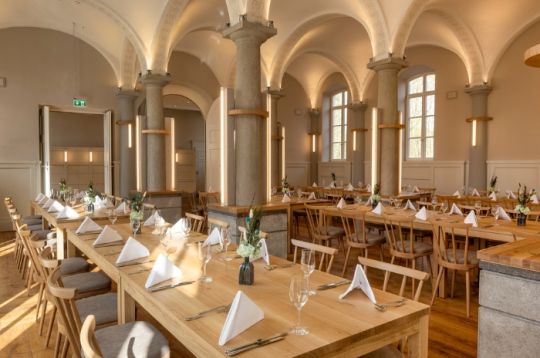 Eleganter Speiseraum mit langen Holztischen, weißen Servietten und gewölbten Decken, in warmes natürliches Licht getaucht.