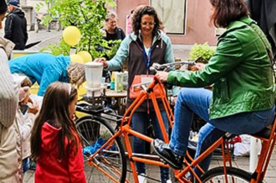 Eine Frau tritt bei einer Veranstaltung in die Pedale eines orangefarbenen Fahrrads, während andere zusehen und ein Kind im roten Mantel interagiert.