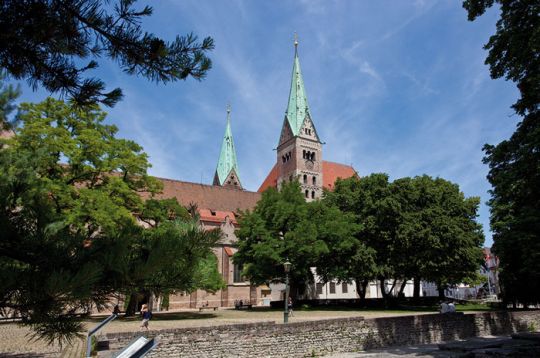 Ansicht des Augsburger Doms mit seinen zwei Türmen unter klarem blauen Himmel, umgeben von grünen Bäumen.