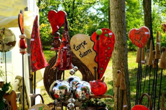 Bunte handgemachte Dekorationen mit Herzformen und 'carpe diem' Schild an einem Marktstand zwischen Bäumen.