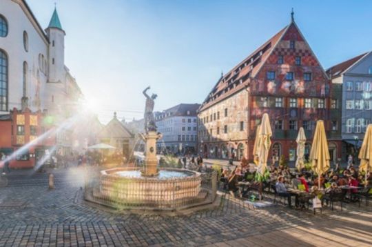 Sonniger Tag am Merkurbrunnen in Augsburg, umgeben von historischen Gebäuden und Menschen, die draußen sitzen.
