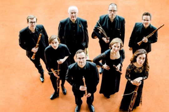 Musiker in eleganter Kleidung mit hölzernen Blasinstrumenten auf einem orangefarbenen Boden.