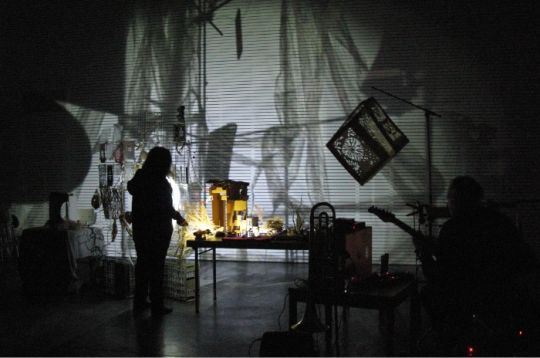 Silhouettierte Figuren mit Gitarren in einem schwach beleuchteten Raum mit Projektionen an der Wand.