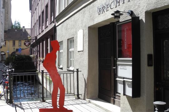 Eingang zum Brechthaus mit roter Statue davor, in einer malerischen Straße gelegen.