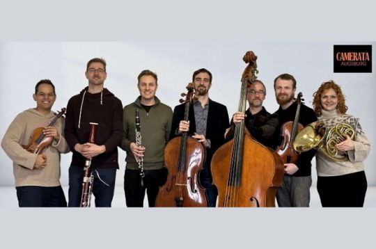 Sieben Musiker mit verschiedenen Instrumenten posieren vor einem hellen Hintergrund.