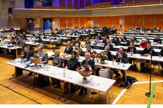 Eine große Versammlung von Personen, die an Tischen in einem Konferenzsaal sitzen.