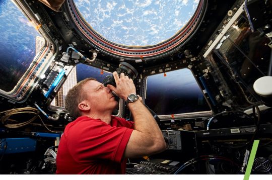 Astronaut fotografiert die Erde aus der ISS-Kuppel, umgeben von Instrumenten, blauer Himmel sichtbar.