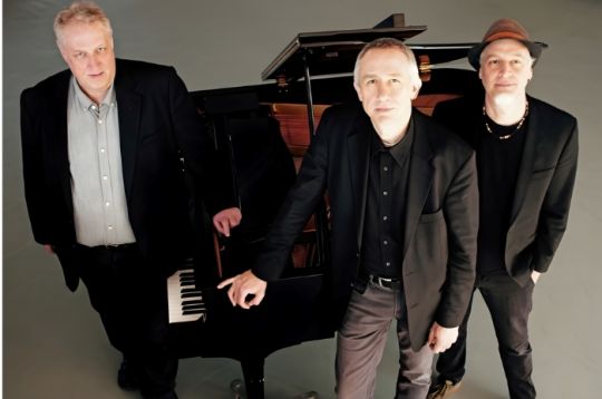 Drei Männer in Anzügen stehen neben einem Flügel und blicken in die Kamera.