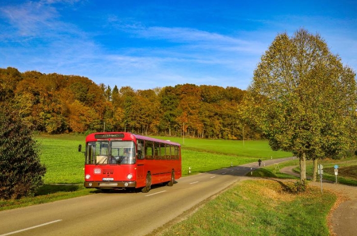 Ein roter Bus fährt auf einer malerischen Straße, mit grünen Feldern und Herbstbäumen im Hintergrund, unter klarem blauem Himmel.