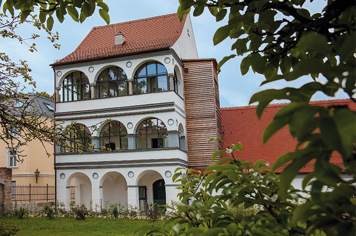 Dreistöckiges historisches Gebäude mit roten Dachziegeln und Bögen, umgeben von grünen Blättern.