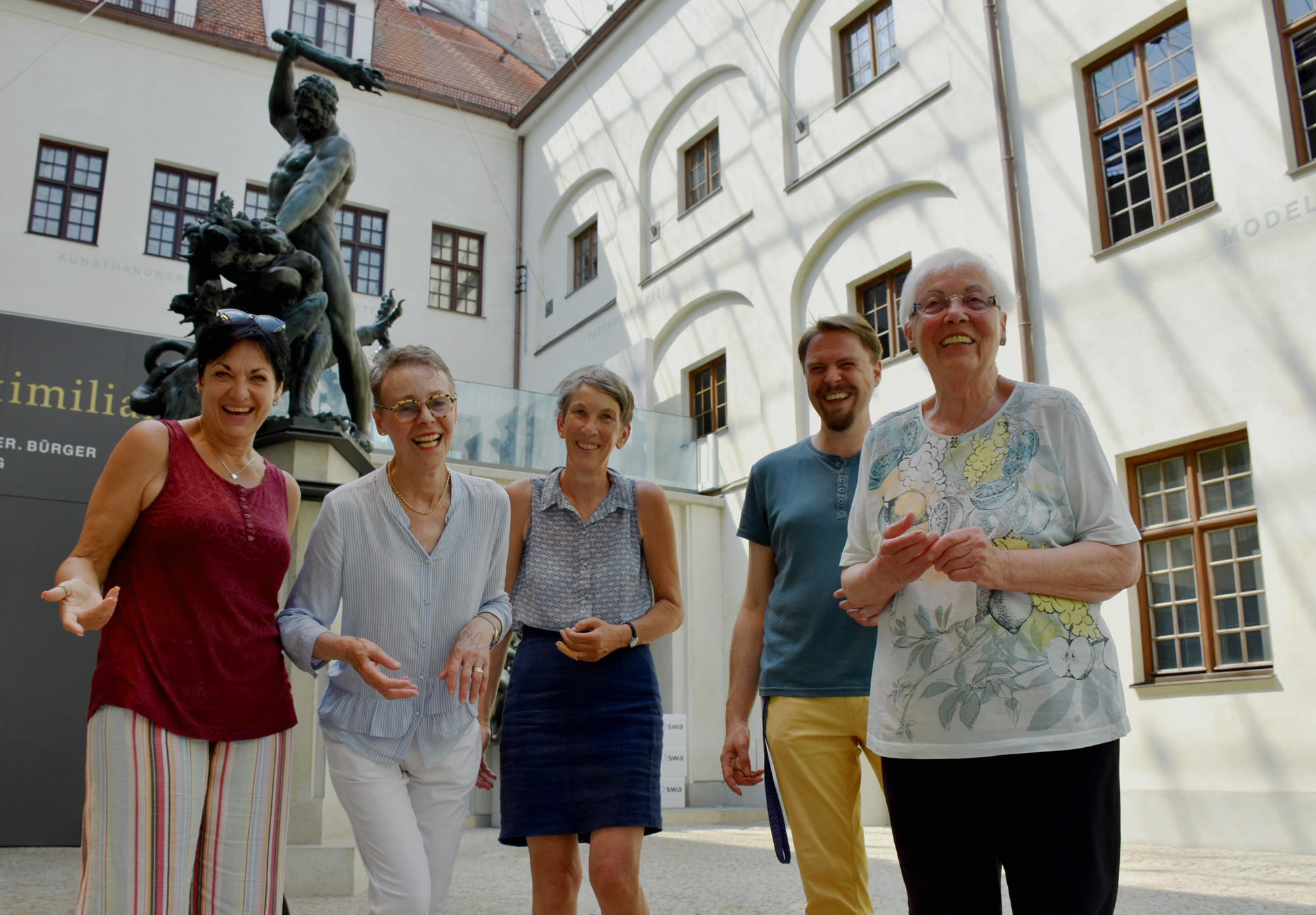 Fünf lächelnde Personen stehen in einem sonnigen Innenhof mit einer Statue im Hintergrund.
