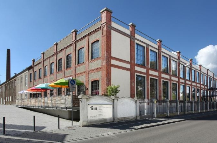 Außenansicht eines roten Ziegelgebäudes mit großen Fenstern, draußen Sitzgelegenheiten unter bunten Schirmen.