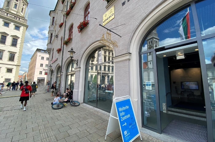 Straßenszene mit UNESCO-Schild und Gebäudeeingang in einer europäischen Stadt, Menschen sitzen und gehen, Fahrräder in der Nähe.