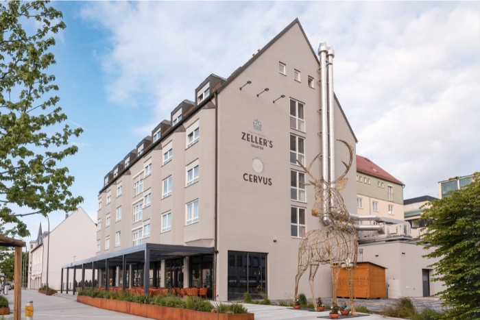 Außenansicht eines modernen Hotels mit einer Hirschskulptur nahe dem Eingang unter blauem Himmel.