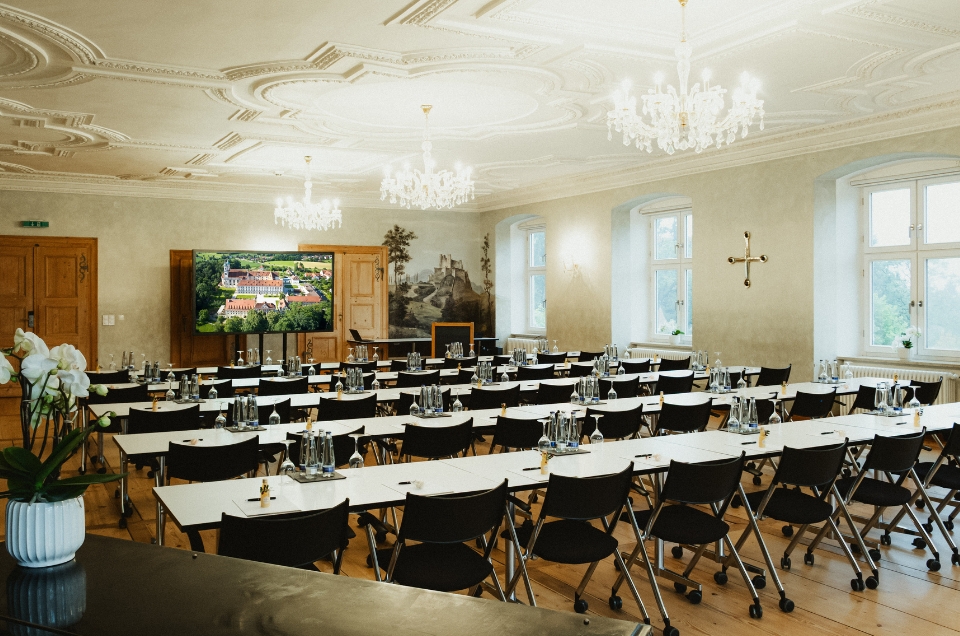 Eleganter Konferenzraum mit Kronleuchtern, Tischen und Stühlen, einem großen Bildschirm und dekorativen Details.